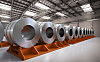 Row of large silver steel coils stacked on orange supports inside a factory.